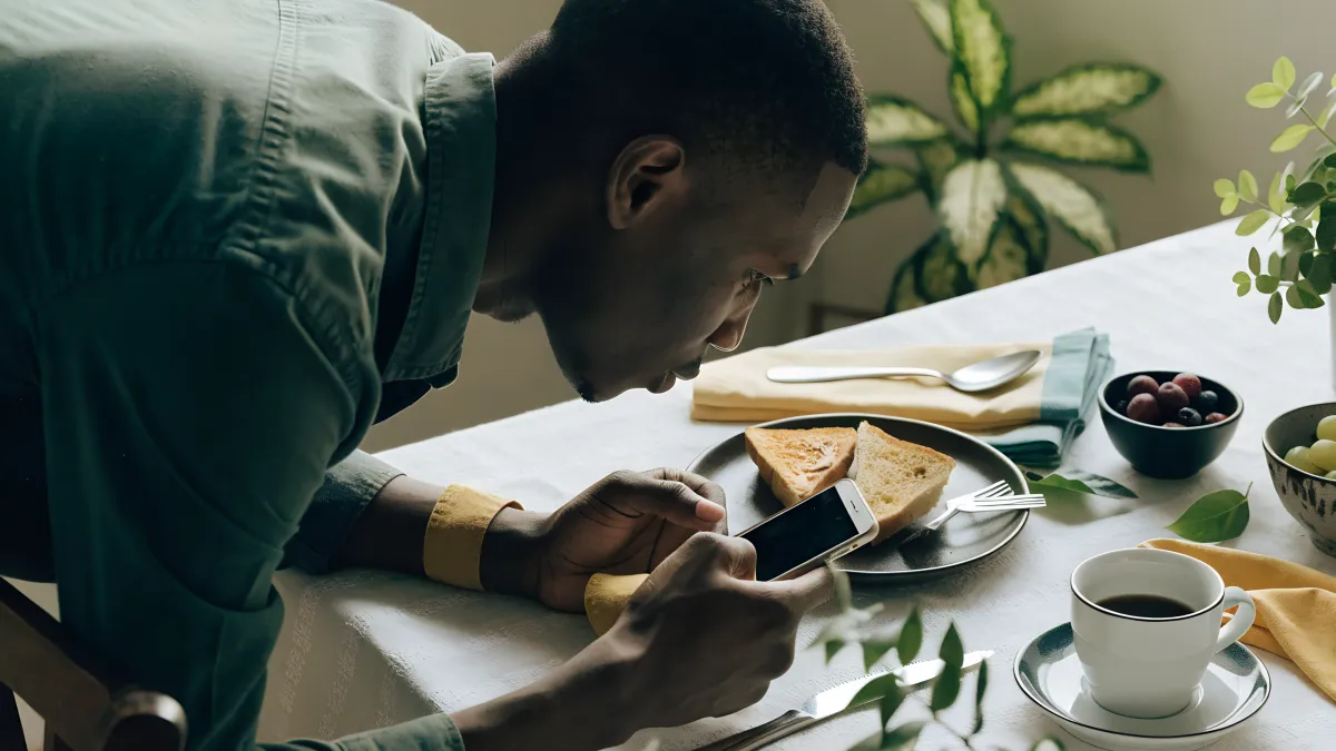 A South African man photographing a styled breakfast flat-lay on a table with his phone, plants around him