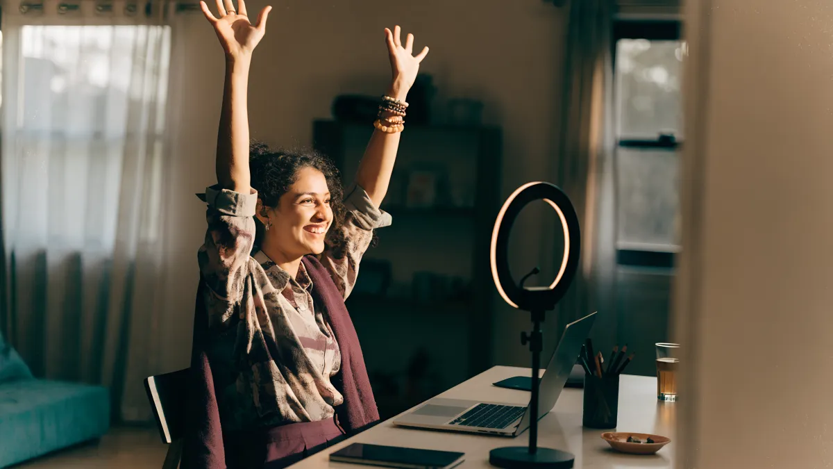 A South African creator celebrating at a desk with a laptop and a ring light, arms raised