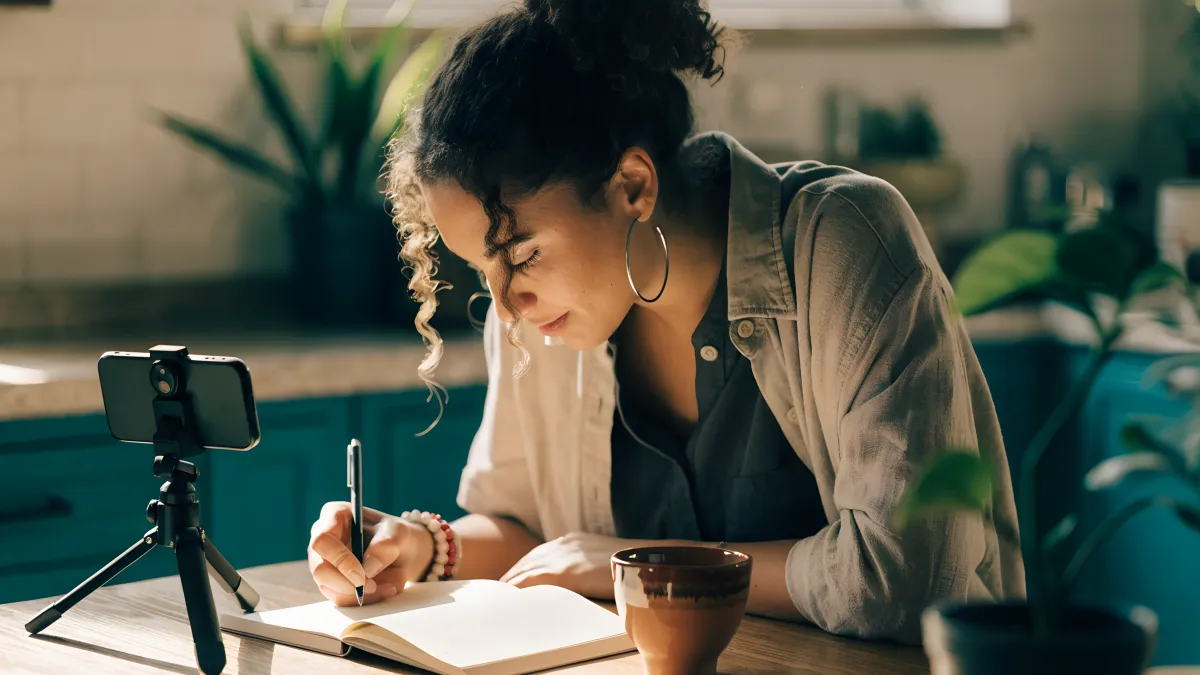 A South African woman in her late twenties at a sunny kitchen table, planning content in a notebook next to her phone on a small tripod, coffee cup, plants